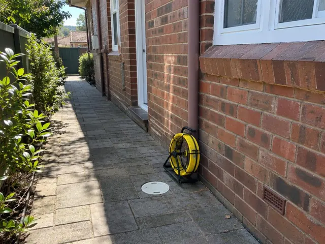 Capped drain inspection cover beside a coiled camera reel on a brick house side path in Pymble