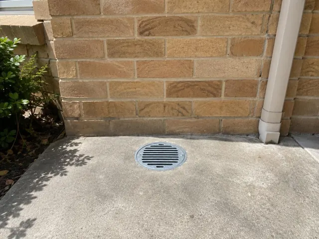 Outdoor gully grate flush in concrete beside brick wall and downpipe in Pymble